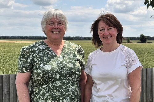 Two people stood in front of fence. One in green pattern dress and one in white t-shirt